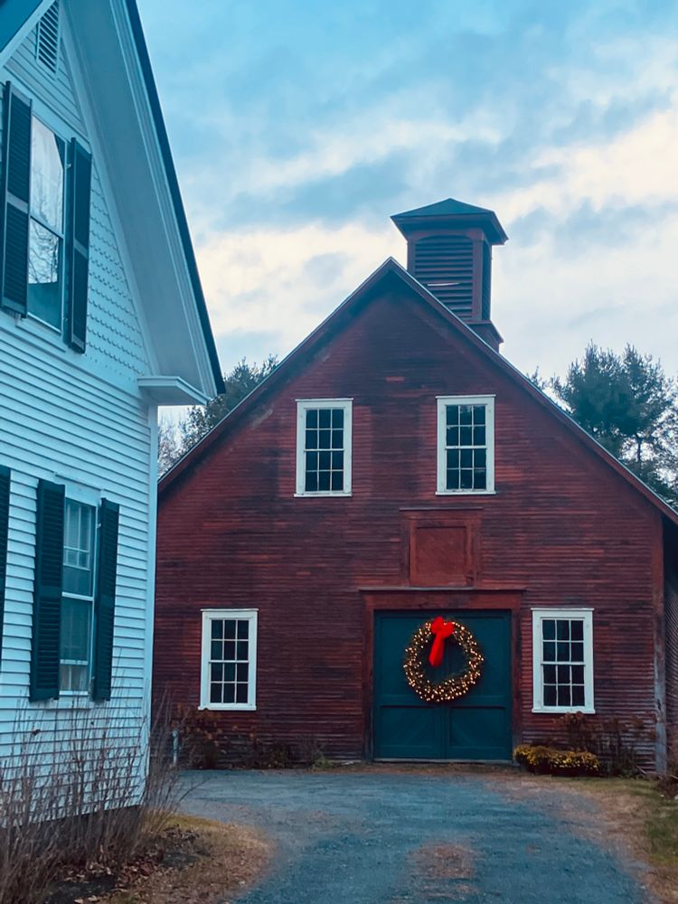 A red barn with a glowing holiday wreath on its blue door, beneath bleak skies 