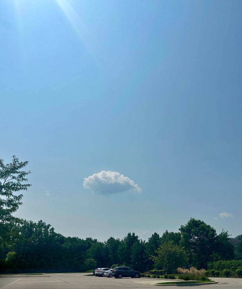 One prominent cumulus cloud is centered above a tree line at the back of a parking lot. The blue sky is hazy and a sun flare is shining down from the upper left corner. 