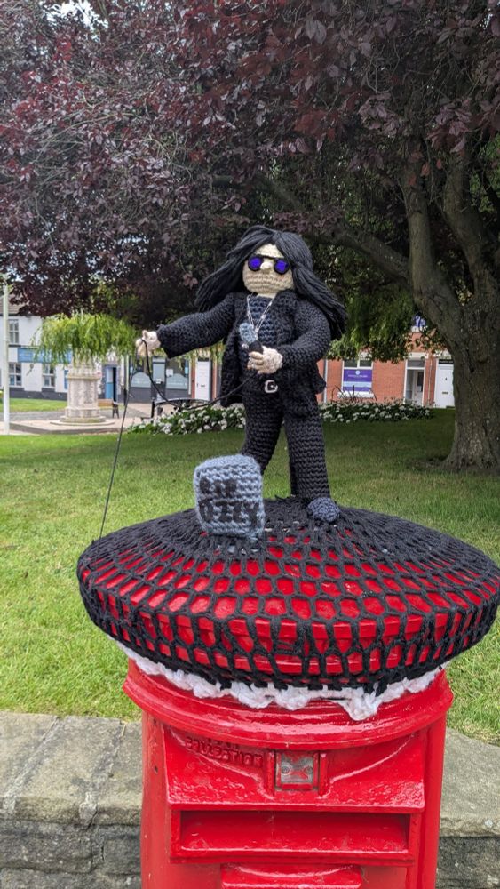 A knitted depiction of Ozzy Osbourne sat atop a post box. A knitted gravestone in front of him says "RIP Ozzy". 