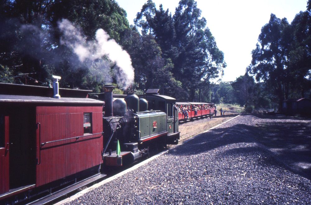 A 2-6-2T narrow gauge steam locomotive in green livery, running bunker-first coupled to a red liveried guards van, sits in a loop as another train, loaded with passengers, many sitting on the window sills and dangling their legs over the side of the carriages, arrives in a platform track to the left.  An amount of white steam or smoke is drifting through the air above the loco, highlighted by the dark green foliage of large trees in the background.