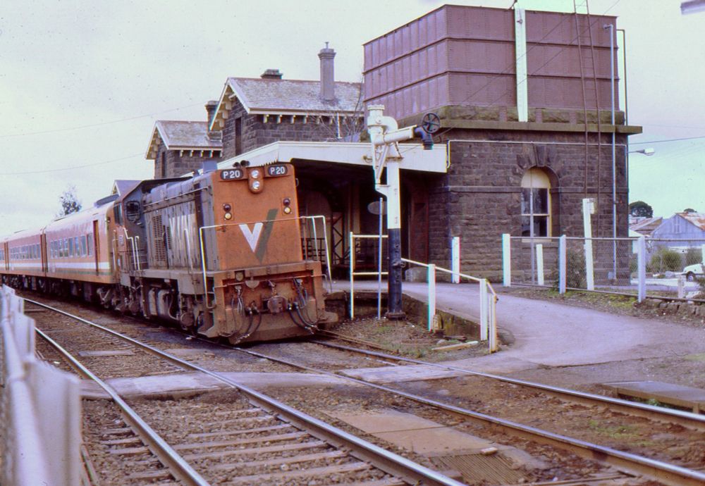 Hood style diesel locomotive P20 in VLine orange and grey livery, long end leading, sits at the head of a three car set of N class passenger carriages also in orange livery.  The train is sitting at a passenger platform in a railway station at which the double track mainline runs between two side platforms.  On the platform at right, behind the train, is a large, two storey bluestone station building with a bluestone-based rectangular water tower.  A steam-era water standpipe is next to the track near the locomotive.  A ground level pedestrian path connecting the two platforms crosses the tracks just in front of the train.