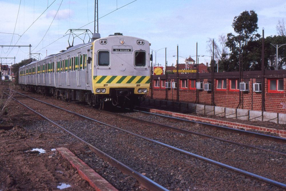 A four car electric multiple unit train painted in light grey livery with green and yellow stripes across the lower front and green and yellow lining at window level along the side, runs towards us along a section of double track railway in a suburban setting.  The train is just passing a construction site at which brick edgings for new low-level tram platforms are partly constructed on each side of the tracks.  To the right of the train, situated very close to the tracks and at a slightly lower level, is the back of a motel.  Further in the distance is a Shell petrol station sign and another sign announcing St Kilda Motor Inn.