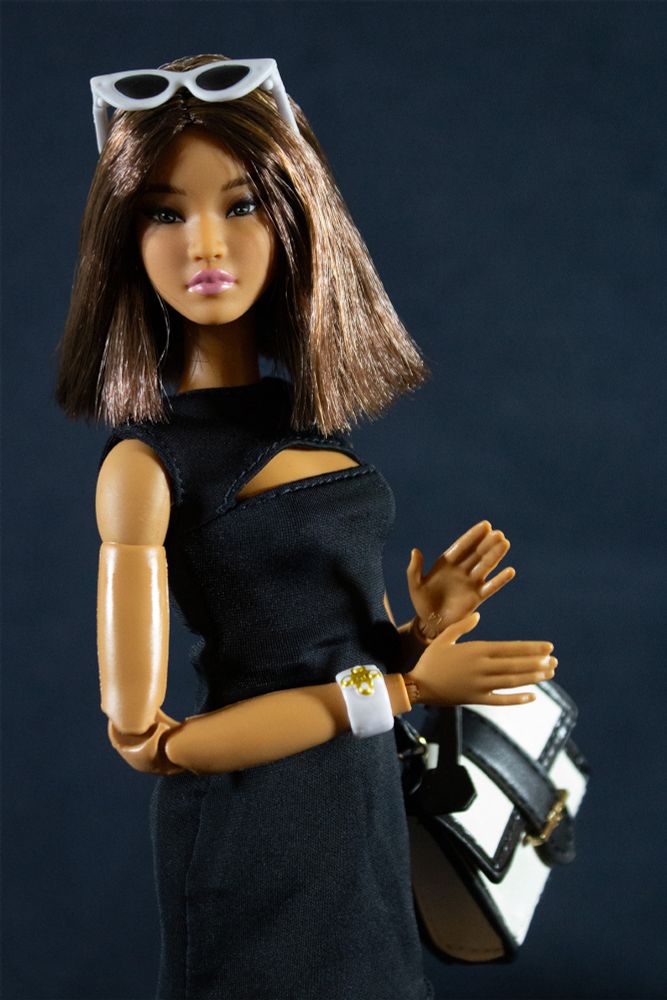 Barbie Basics doll with the Kit face sculpt, with subtle pink makeup, on an Original Made To Move body in a medium deep tan skintone. She has shoulder-length dark brown hair cut in a blunt angled bob, and has white sunglasses with black lenses atop her head. She wears a sleeveless black minidress with a boat neckline and a cutout that forms an elongated crescent shape across her upper chest. She also wears a white and gold cuff bracelet and carries a black and white handbag with a gold buckle.