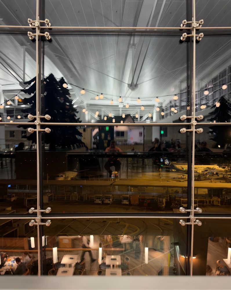 Picture of an airport window looking out the tarmac at night. The interior of a too well lit “bar” and its weirdly plastic decor reflects in the window. In the center a lone male figure sitting and wearing a sweatshirt that is hi-vis orange is in the center of the reflection. It is a self portrait. 