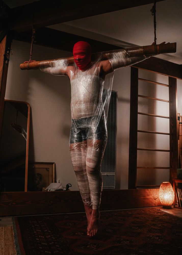 A person is suspended from a bamboo beam hanging like Jesus on the cross. Their head is coved in red tape, their body bound in white gauze and pallet wrap