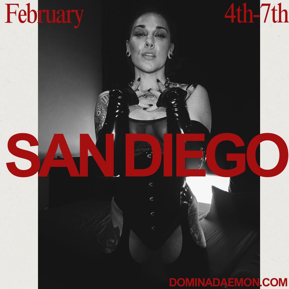 Domina Daemon dominatrix is standing in black latex behind an advertisement for a San Diego tour. 