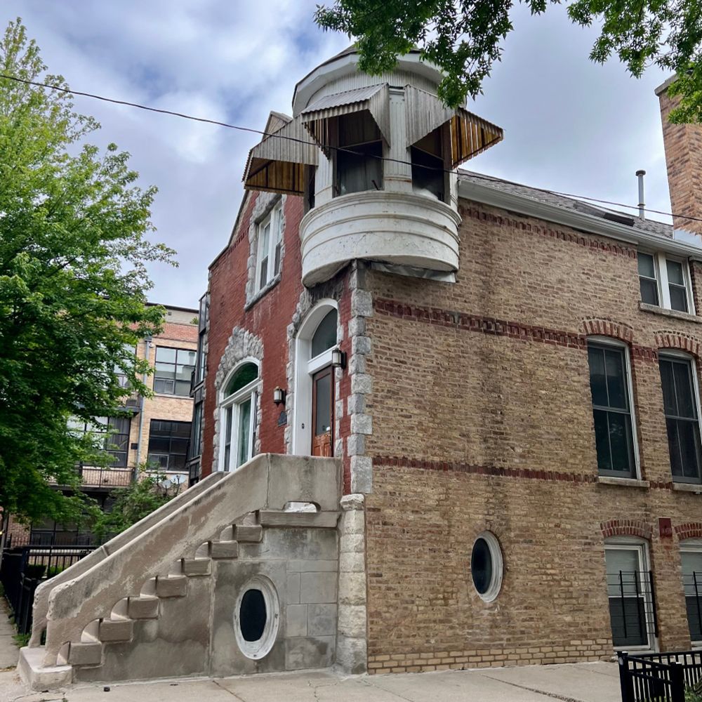 Brick cottage with stone quoins, stone window trim, and stone steps and railings. There is a small round turret at the corner of the second floor with a conical roof. The windows on the turret are protected by wooden awnings that are a bit too big and sort of comical. There is a creepy oval shaped window in the side of the concrete/stone? steps that opens on to who-knows-what.  There is also another random oval window in the first floor brick wall. The whole thing screams fairy tale.
