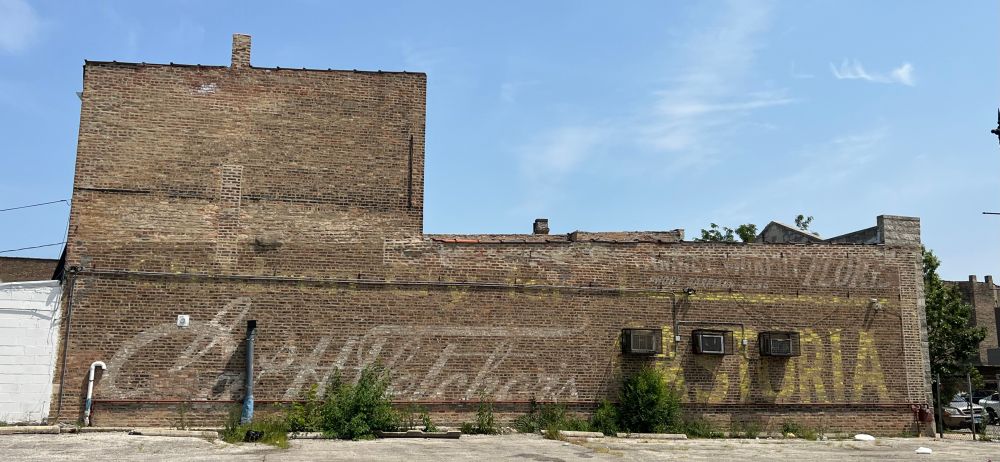 The side of a building that is 2 stories on the left and a single story on the right. It is built of Chicago common brick. Painted on the side in yellow letters is "Children Cry for" then in white script "Chas. H. Fletcher's" and again in yellow all caps "Castoria." A few scrubby bushes obscure a bit of the writing.