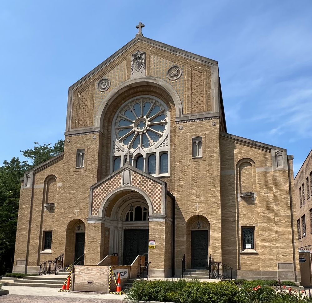 Large symmetrical church of buff brick. The Chicago Historical Resources survey calls this style Romanesque Revival. The entrance projects out from the center of the church in the form of a brick archway leading to the door. The top of the archway is a checkerboard brick pattern of bluff and cream tiles with a stone border. Above the entrance is a large round wheel window decorated with stone tracery. And above that is more checkerboard tile, stone carvings of religious symbols, and a stone cross at the very top. One either side of the main entrance are two other doors set in arched openings.