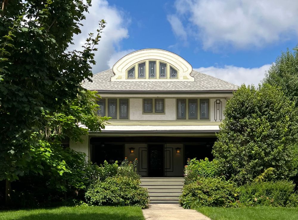 Beautiful Prairie style stucco home with an open porch, leaded glass windows, and a curved dormer at the front of the hipped roof. Trees and shrubbery cover both sides of the building.