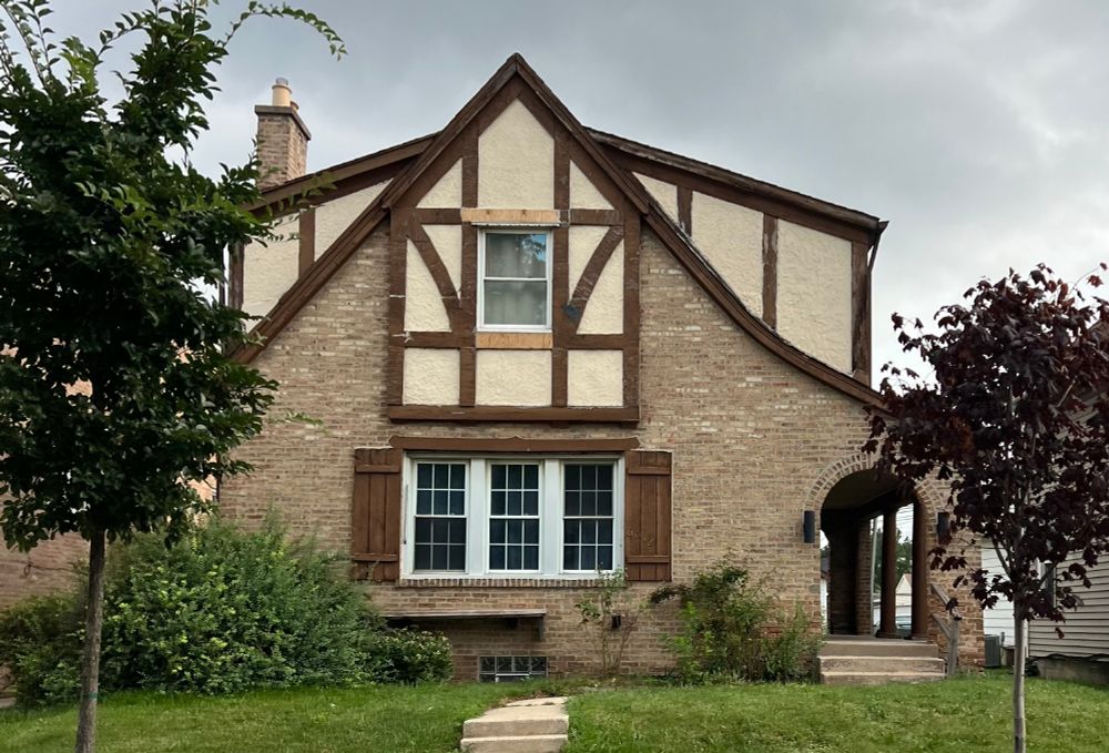 Tudor-style two-story home of tan brick with half timbering on the second story. There is a steep gable roof that curves up a bit on the right side to make room for an arched passageway underneath it. Two half timbered dormers stick out like ears on either side of the gable peak. The half-timbering consists of off white stucco with brown beams crossing it. A couple of the beams have been replaced and are unpainted. three multi-paned windows are centered on the first floor, below the half timbering. They have brown wooden shutters on each side. 