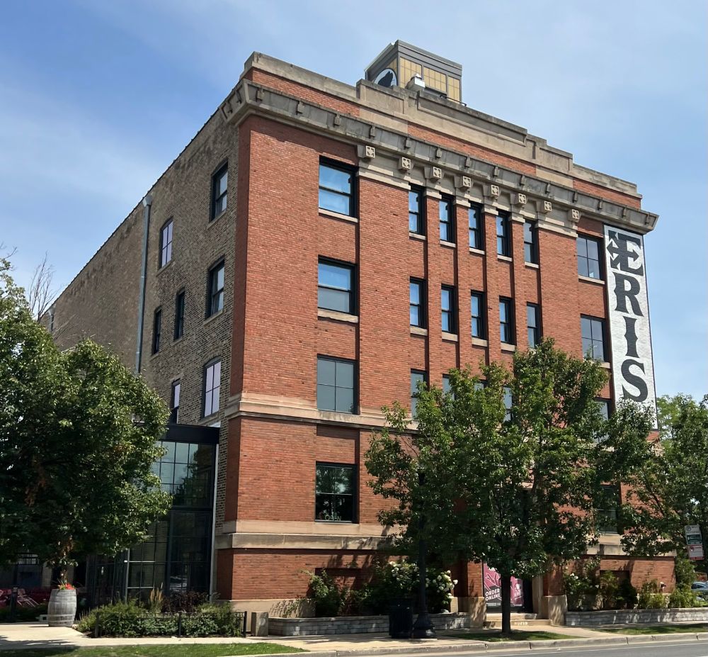 Four-story+ brick building with red face brick and Chicago common brick on the side. The facade has 5 narrow vertical windows closely spaced on floors 3-5 as well as larger windows on each side of all floors. The windows have stone sills and there is stone decoration across the top of the building.