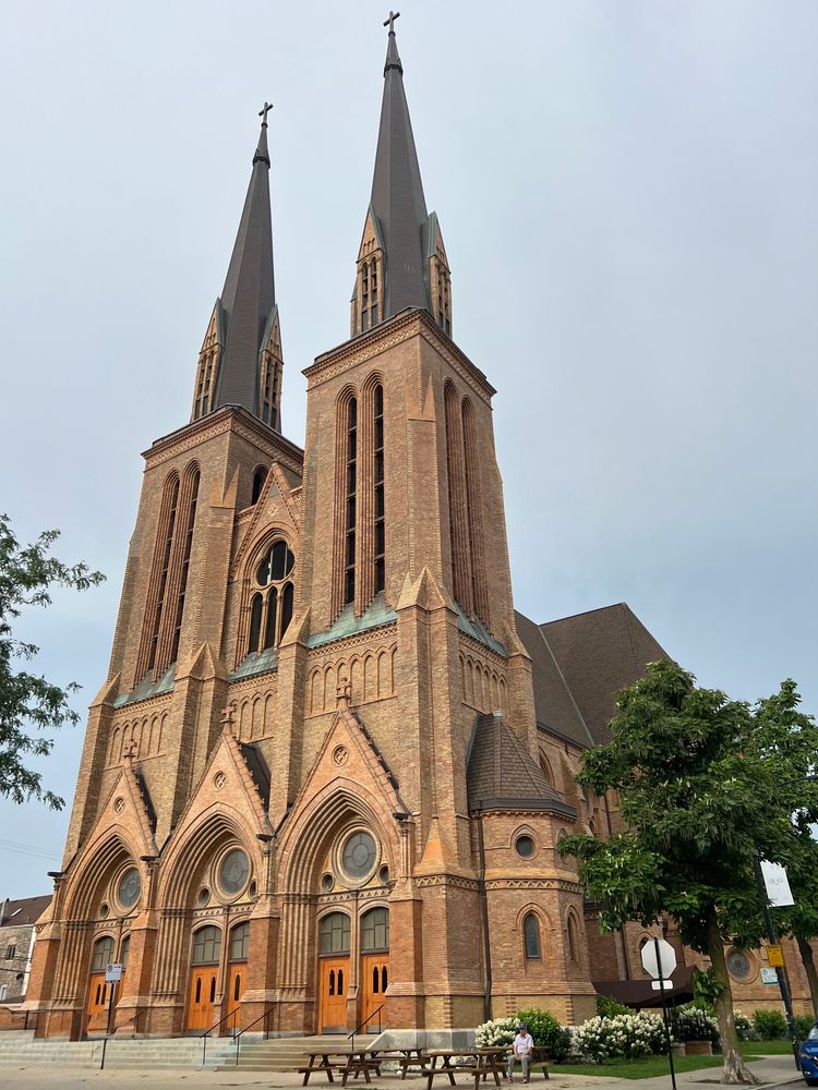 Brick Gothic church with two soaring spires topped with crossed. There are three entrances on the front, each with double doors. Above the entrances are multilayered brick arches, round windows, and brick pediments topped with terra cotta crosses. The brick is buff-colored and really glows in the sun.