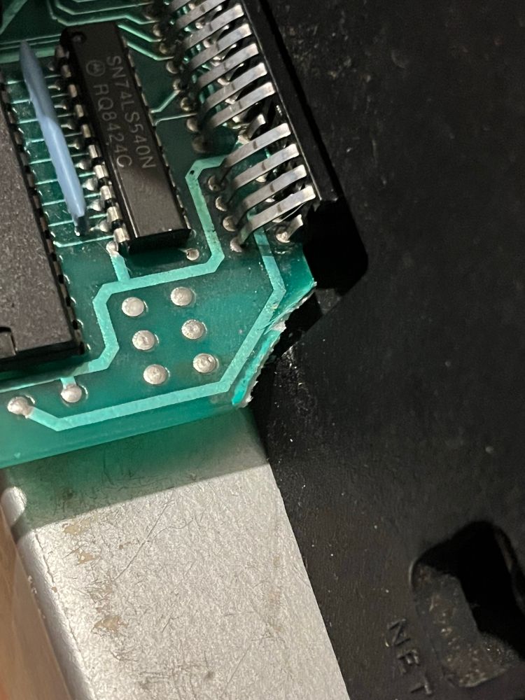 Close-up image of a circuit board featuring a black chip labeled "SN74LS540N," with visible solder connections and traces on a green substrate. The circuit board is positioned against a metallic surface. A corner of the board has been removed