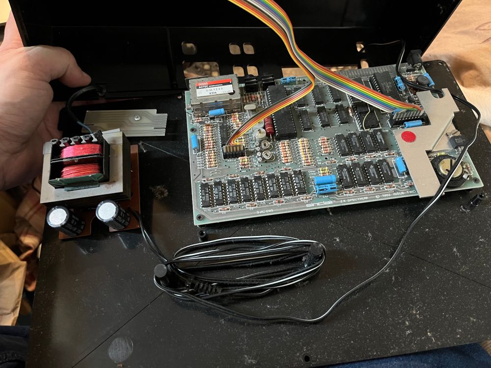 An opened electronic device, featuring a circuit board with various components and connectors. A transformer and capacitors are visible, along with connecting wires. The background shows part of a hand and a textured surface.