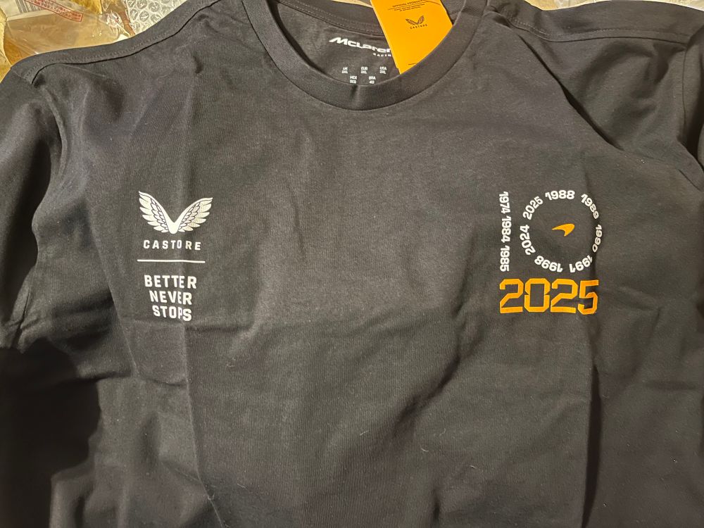 A black t-shirt with white and orange text, the collar has a McLaren logo with a 2025 logo on the breast
