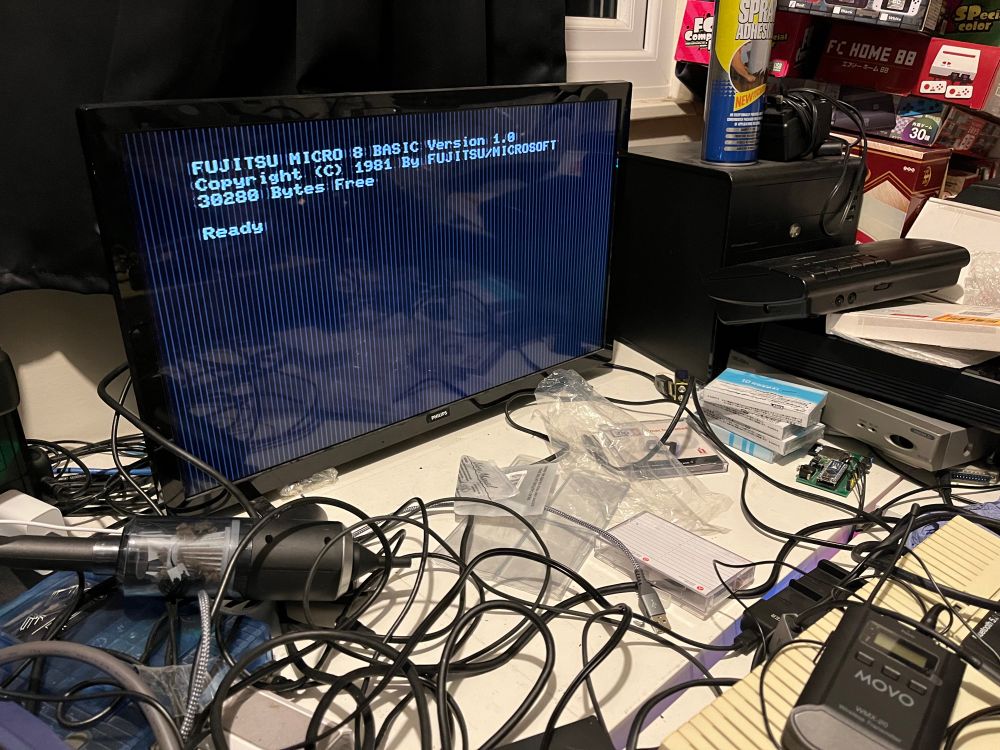 A cluttered desk featuring a monitor displaying a BASIC programming prompt for a Fujitsu Micro 8 computer from 1981. Various cables and electronic devices are scattered across the surface, including a black computer tower, a microphone, and miscellaneous packaging.