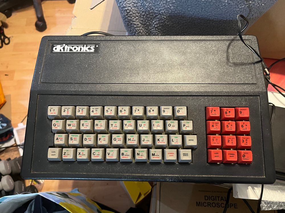 An old computing device featuring a rectangular keyboard with a mix of gray and red keys. The device has a logo on the top that says DK Tronics