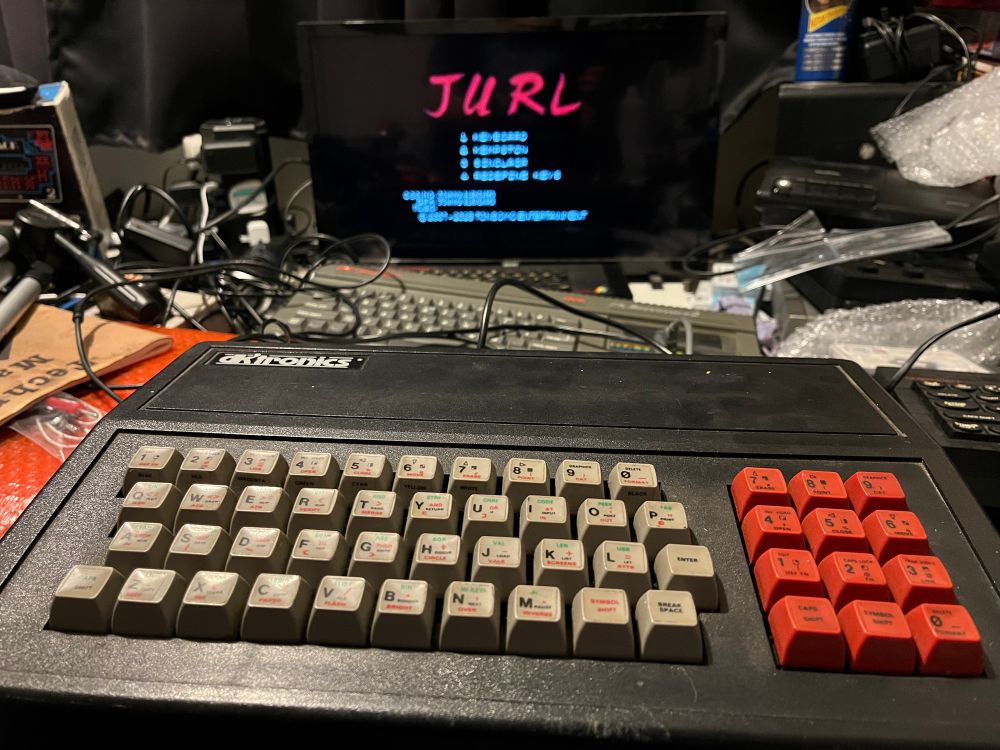 A computer with grey and red keys, the logo says dk tronics. It’s connected to a screen which has the title for a game called Jurl