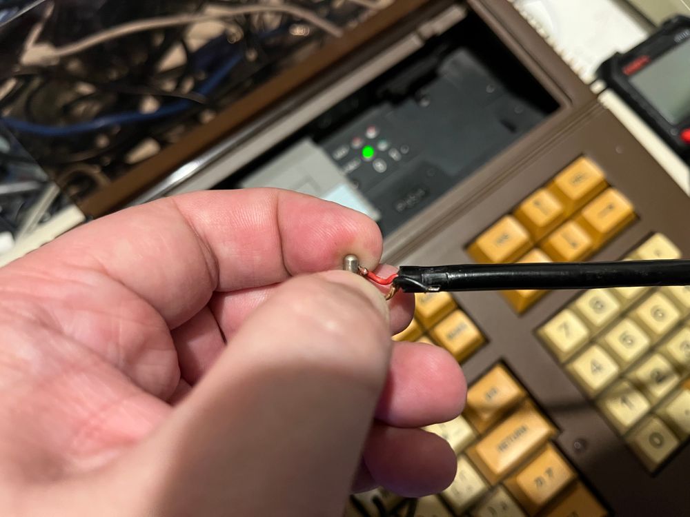 A hand holds a wire with a red connector, while a retro-looking device with yellow keys is partially visible in the background. 