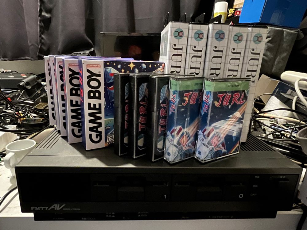 A collection of retro video game cartridges, including Game Boy boxes and cassette-style games, displayed on top of a black computer. Various cables and electronic items are visible in the background.