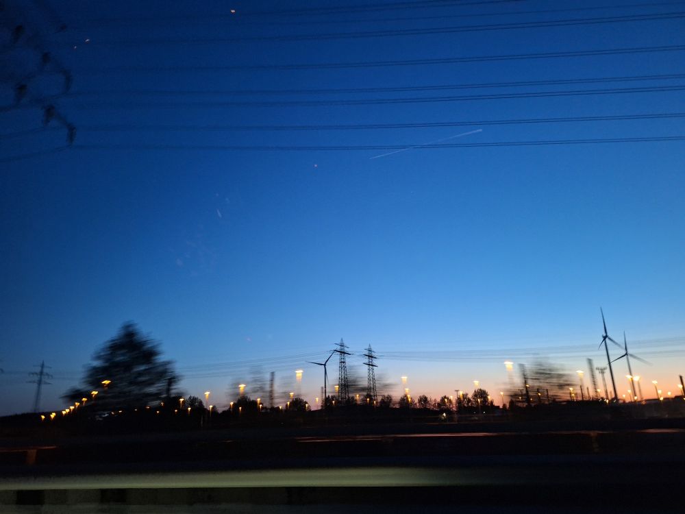 Foto aus dem bewegten Auto. Die Sonne ist untergegangen und es ist nur noch ein dünner Streifen in orangegelbem Licht. 
Am oberen Bildrand sind Stromleitungen. In der Bildmitte weiter weg sind Windkraftwerke, Beleuchtungem und weiter Stromleitungem verschommen sichtbar.