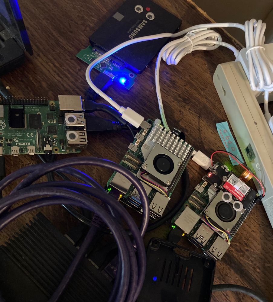 three raspberry pis and a 2.5" ssd on a wooden surface, with a dark purple coiled cable messily in the foreground. two of the raspis have google eyes and so does the ssd, but the middle pi does not (yet!)