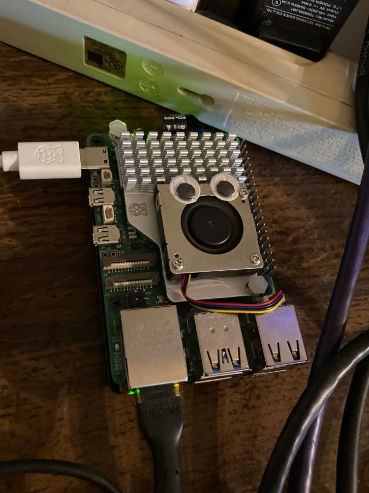 a raspberry pi 5 with active cooler on top with googly eyes on its fan

a white raspberry pi-branded power cable is connected to its USB-C port, and a black networking cable connected with green and amber LEDs lit. it's on a dark wood surface with part of a power strip behind. several networking cables are messily crossing the edge of the frame.