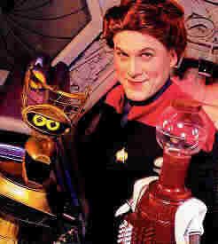 Crow T. Robot and Tom Servo flank Mike Nelson, the latter of whom is dressed in a Starfleet uniform and wig to look like Kate Mulgrew as Kathryn Janeway.