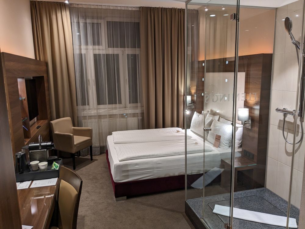 Bild eines sehr luxuriösen Hotel Zimmers mit Doppelbett und einer gläsernen Dusche, ungewöhnlich mittig im Raum.