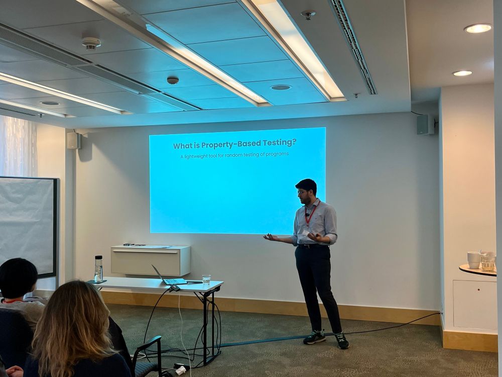 A person presents at a conference. Some audience members are visible in the left corner of the image.
