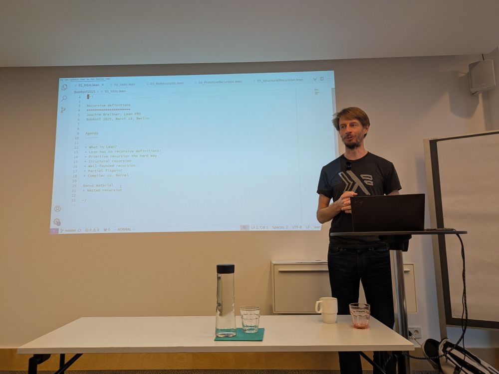 A person presents at a conference. They are wearing a t-shirt with a lambda symbol on it.