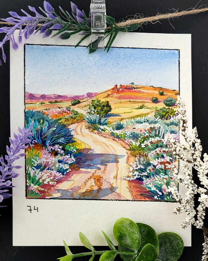Square watercolor framed with a black line on a rectangular, portrait-oriented paper. A sandy path winds through an arid landscape with sparse shrubs and rocky hills. White and pink blossoms are scattered around. The warm earth tones contrast with the cool blues of the sky. Number 74 is written in the bottom left corner.