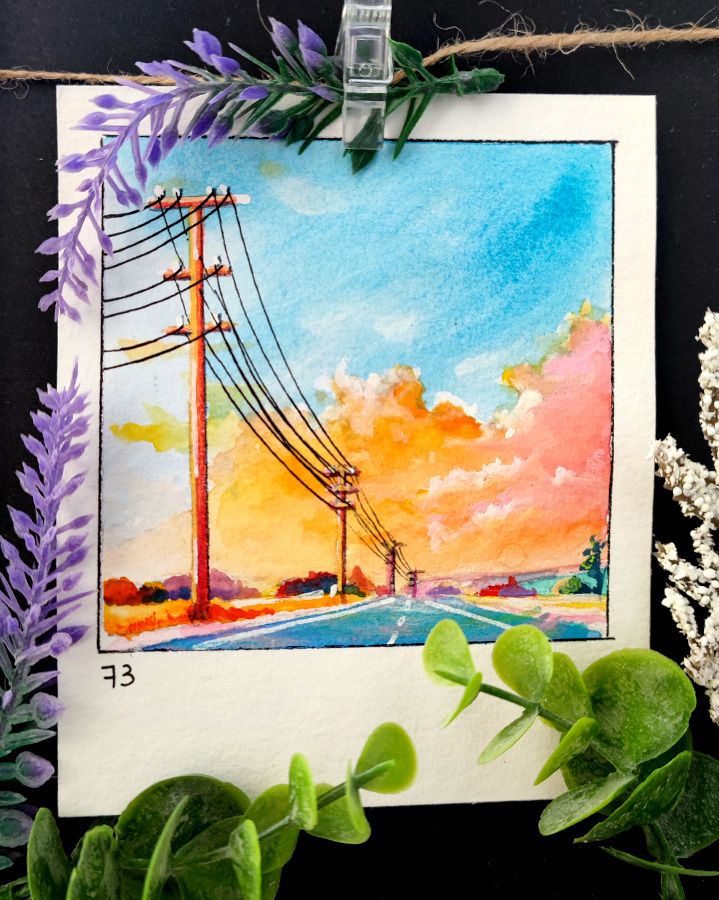 Square watercolor framed with a black line on a rectangular, portrait-oriented paper.
An endless road stretches into the distance, accompanied by towering power poles whose cables cut through the scene. The sky glows in warm orange and pink hues as the last sunlight illuminates the drifting clouds. The landscape is bathed in soft shadows, while the deep blue asphalt contrasts with the vibrant evening sky. Number 73 is written in the bottom left corner.