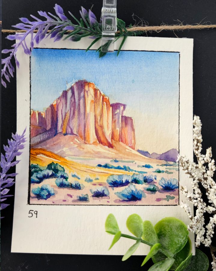 Square watercolor framed with a black line on a rectangular, portrait-oriented paper. A towering rock formation rises into the clear sky, surrounded by a desert landscape with violet-blue shrubs. The warm light enhances the vibrant colors of the cliff face. Number 59 is written in the bottom left corner.