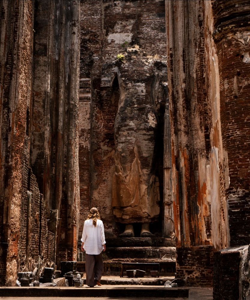A woman with long, light brown hair, dressed in a loose white shirt and gray pants, is walking barefoot toward a towering ancient brick statue in Polonnaruwa, Sri Lanka. The statue, missing its head, is situated within the weathered ruins of a temple with intricate, textured brick walls showing signs of age and erosion. The lighting highlights the earthy tones of the brickwork, casting a serene and contemplative atmosphere around the historic site. Broken stone artifacts and debris are scattered along the pathway, emphasizing the sense of antiquity.
