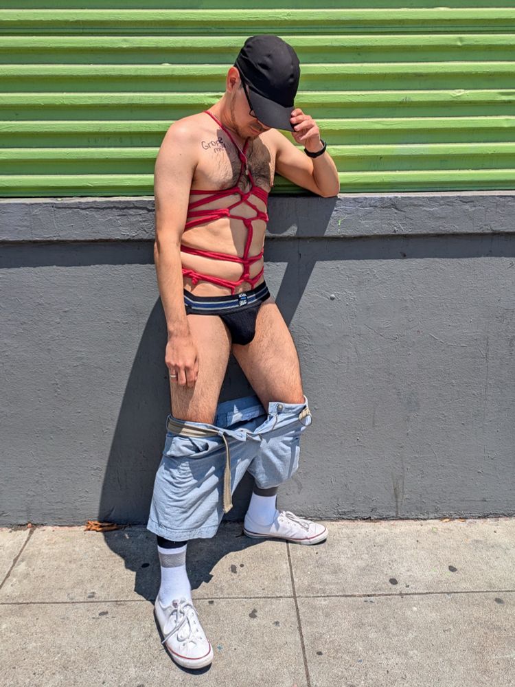 The same man as before, his shirt removed and his shorts lowered down to his knees, revealing more of the red rope around his torso and chest. His underwear is a black jockstrap. He has GROPE ME written around his right shoulder.