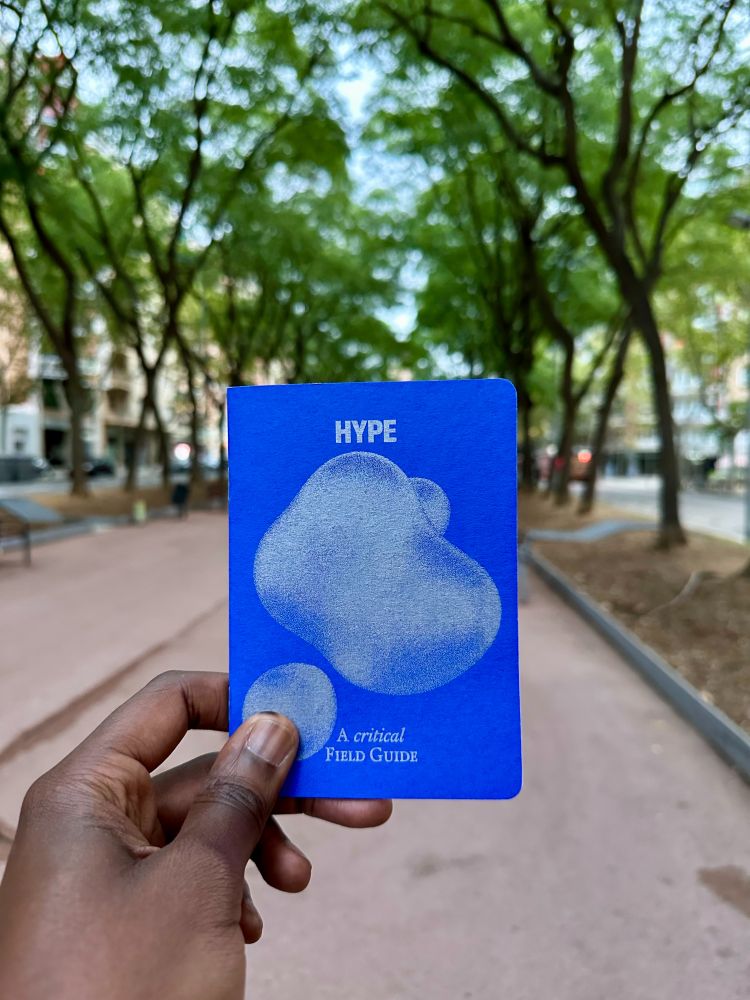 A POV shot showing a hand holding a small passport sized self-published booklet called HYPE: A critical field guide by the German design researcher Johannes Klingebiel.