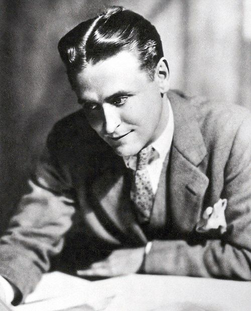 Black and white photo of F. Scott Fitzgerald with an eager & interested look on his face, leaning on a desk, looking down at sheets of paper.