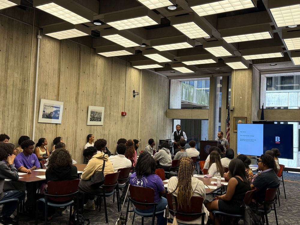 Wilmer Quinoñes, Councilor Santana's Director of Youth & Community Engagement and Jordan Frias from Councilor Weber's Office lead a discussion with youth from BSAC & Sociedad Latina seated at tables in a large room in Boston City Hall