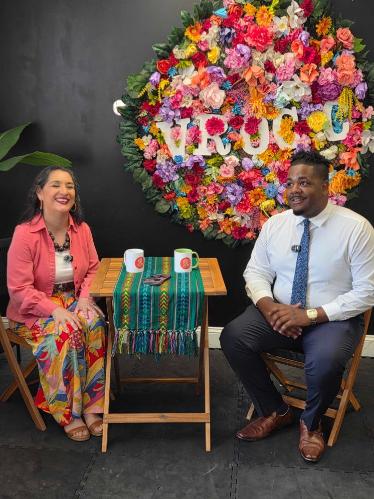 Councilors Santana seated with Veronica Robles on the interview stage before today's interview with a large multicolored floral wall adornment in the background