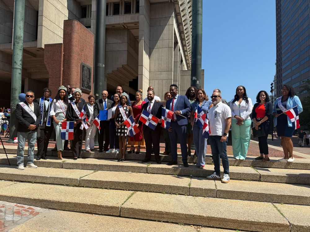 A group of community advocates and elected officials and staff standing on City Hall Plaza with Dominican Flags during the Dominican Flag raising. 