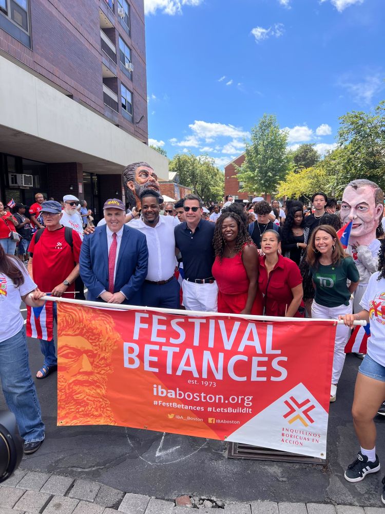 A group photo of Councilor Santana with fellow elected officals, Councilors Flynn, Fitzgerald, Louijeune, and Mejia with the Festival Betances Parade banner with large paper mache heads (cabezudos) in the background 