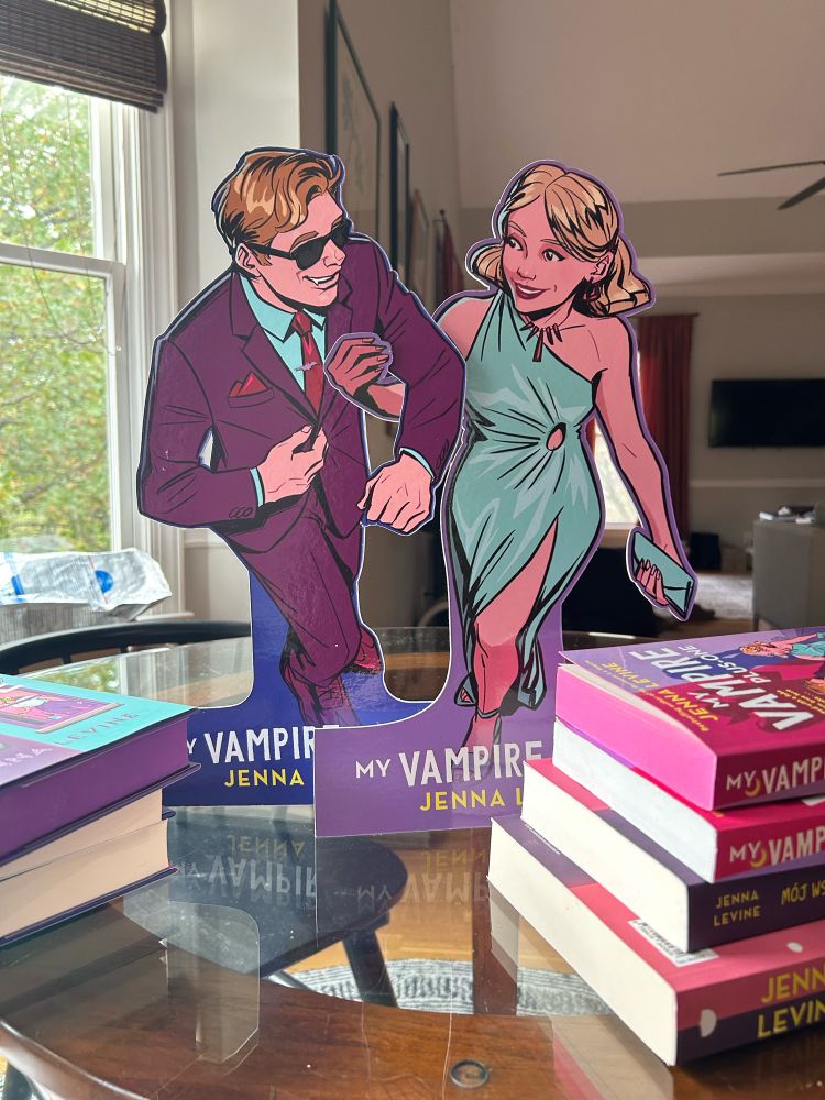 Cardboard cutouts of Reggie and Amelia from MY VAMPIRE PLUS-ONE on a glass-top table. Stacks of different editions of MY ROOMMATE IS A VAMPIRE and MY VAMPIRE PLUS-ONE are also in the picture