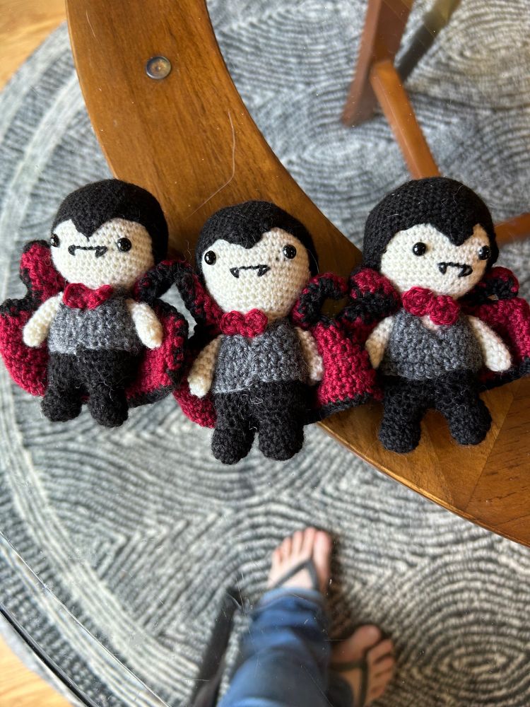 Three vampire amigurumi on a glass-topped table. Gray and white carpet and photographers flip-flops are visible 
