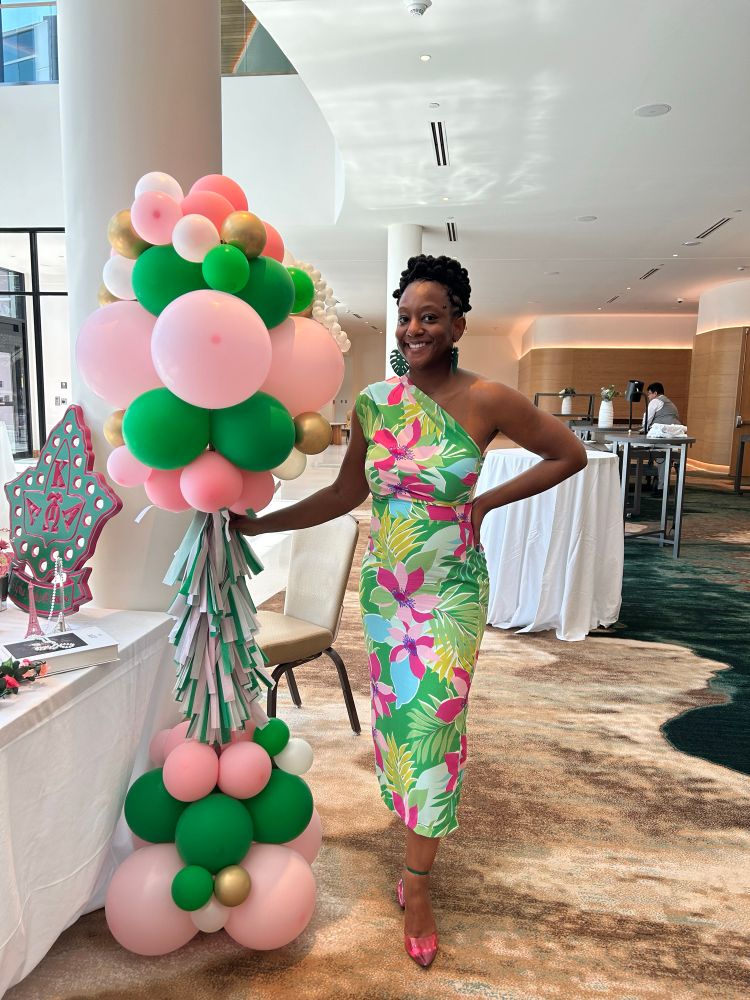 Picture of Black woman in a pink, green, blue dress with pink and green balloons. 