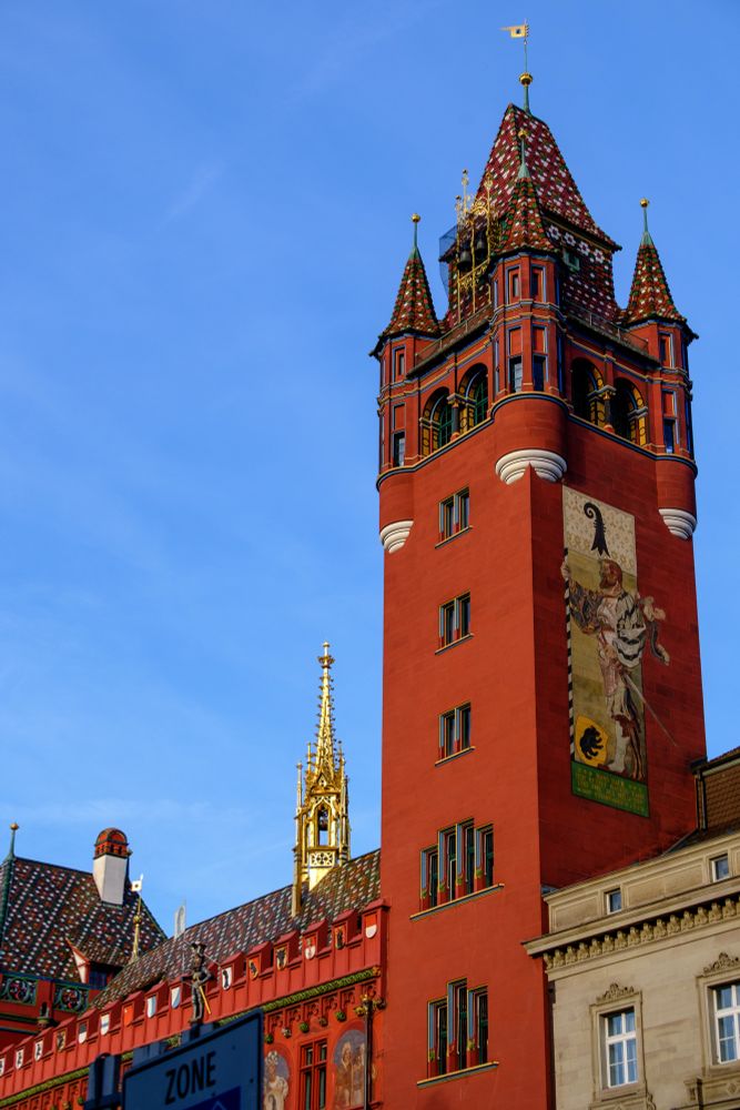 Der Turm des farbenprächtig verzierten roten Rathauses in Basel. Auf dem Dach an vergoldeter Spitzturm. Blauer Himmel. 