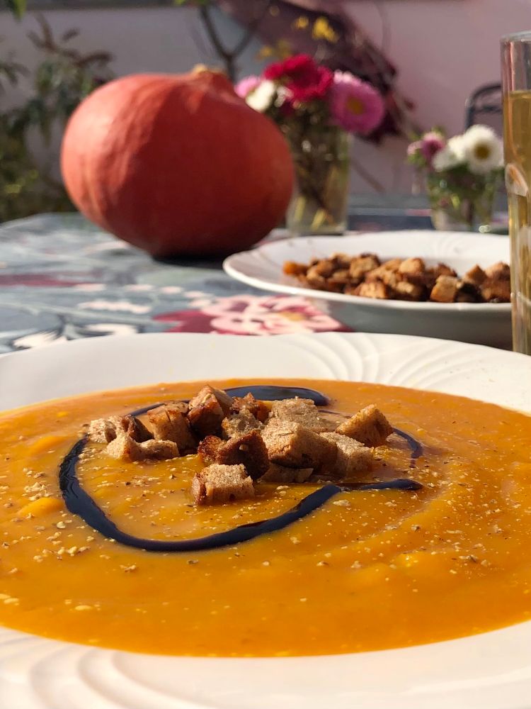 Kürbissuppe mit gerösteten Brot und einem Spritzer Balsamico. Im Hintergrund liegt ein kleiner Kürbis auf den Tisch. 