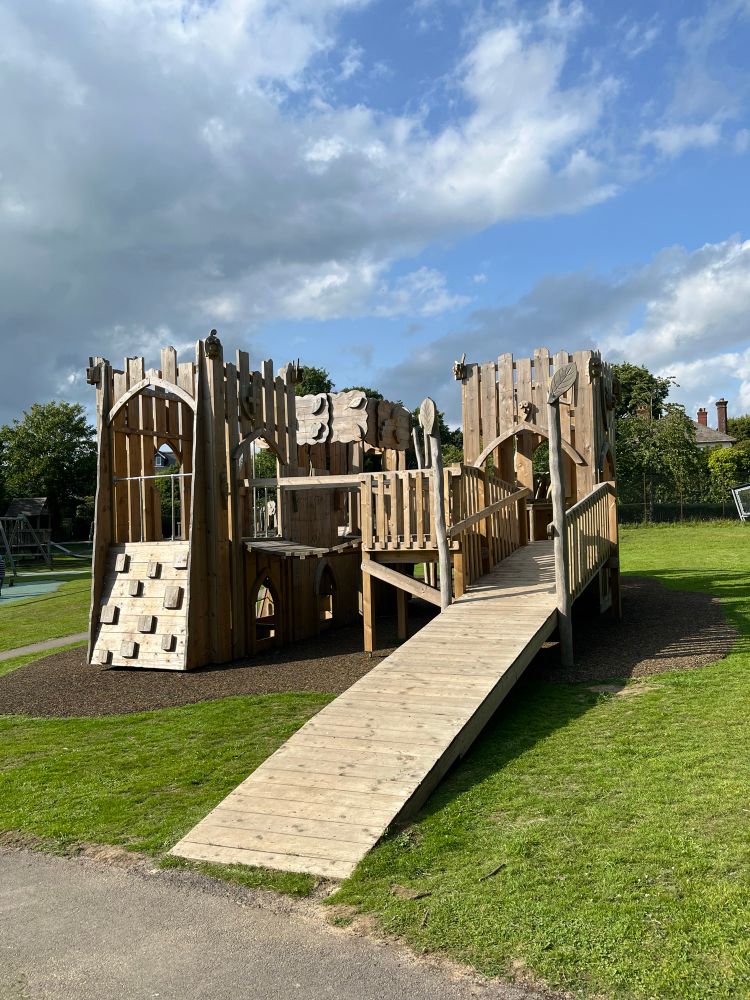 Eine große Kletter- und Spielburg aus Holz auf einem Kinderspielplatz. Eine große Rampe führt in die Burg und kann so auch so Menschen im Rollstuhl oder mit Gehbehinderung genutzt werden. 