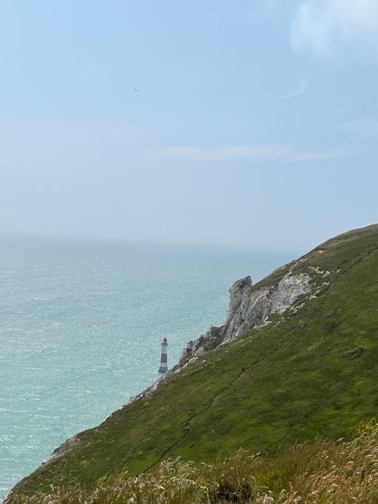Eine Steilküste, hellblaues Meer und in der Ferne ein kleiner rot-weißer Leuchtturm, Beachy Head. 